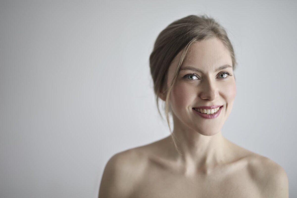 Close-up portrait of a smiling blonde woman against a gray background, embodying elegance and beauty.