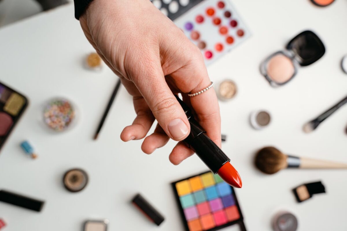A hand holding a red lipstick surrounded by various makeup products on a light background.